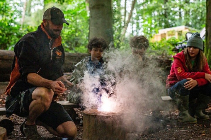 People gathered around a fire pit in a forest, with smoke rising.