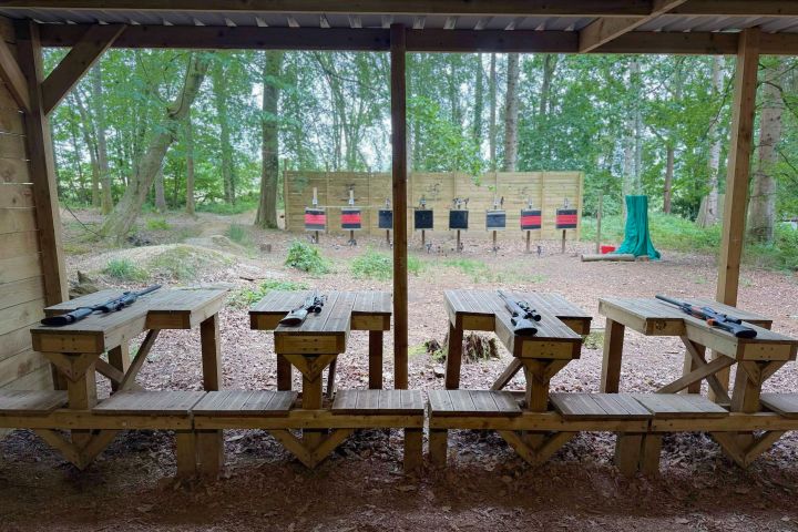 Covered shooting range with rifles on benches, aiming at targets in a wooded area.