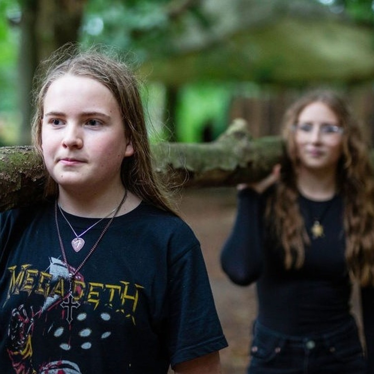 Three teenagers carry a large branch together in a wooded area.