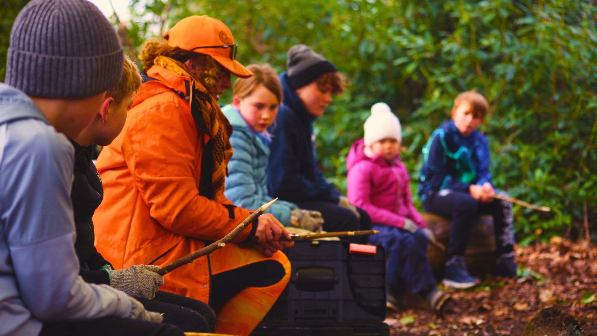 Group of children with a guide in orange sitting in a forest holding sticks.