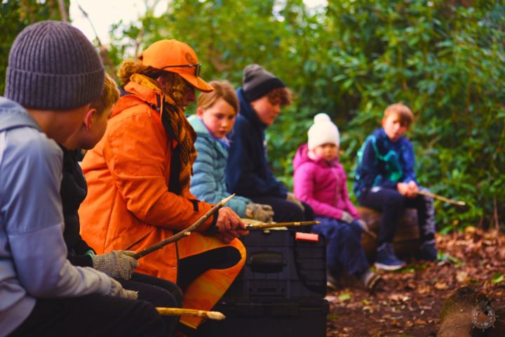 Group of children with a guide in orange sitting in a forest holding sticks.