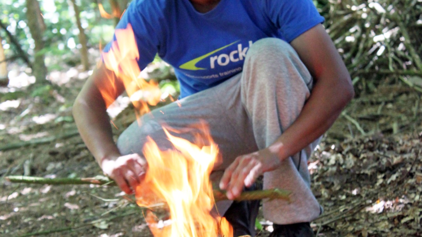 Person building a campfire in the woods, wearing an orange cap and blue shirt.