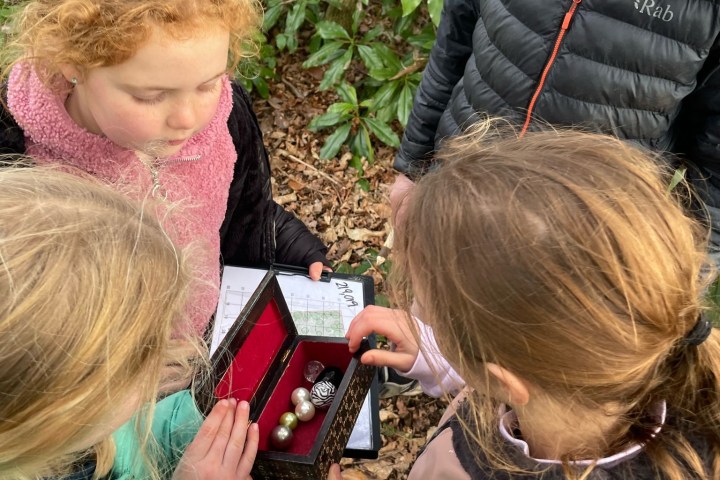 Children gathered around a small open box with trinkets in a leafy outdoor area.