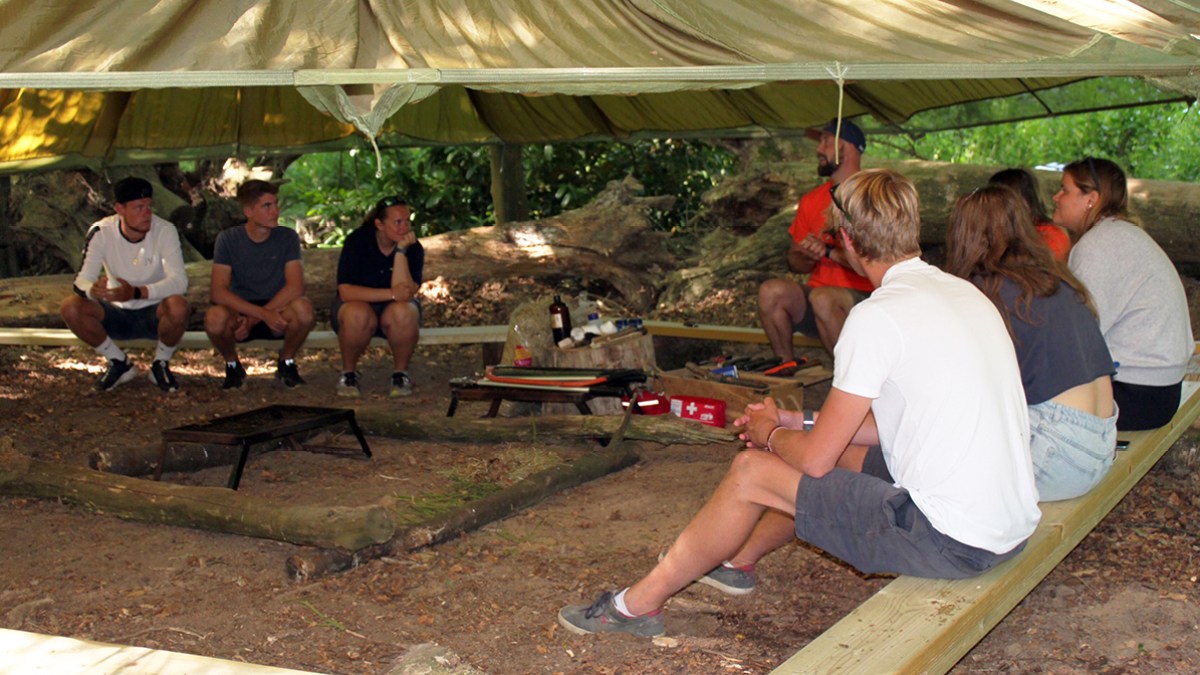 Group of people sitting under a tarp shelter in the woods, engaged in conversation.