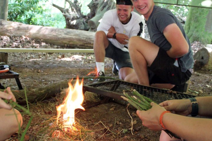 Two people smiling near a campfire holding sticks, outdoors in a forest setting.