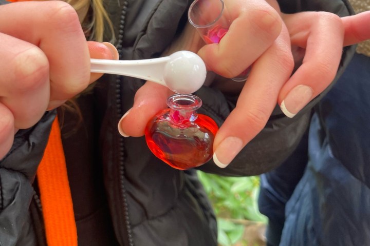 Person filling a small red glass bottle with funnel and scoop outdoors.