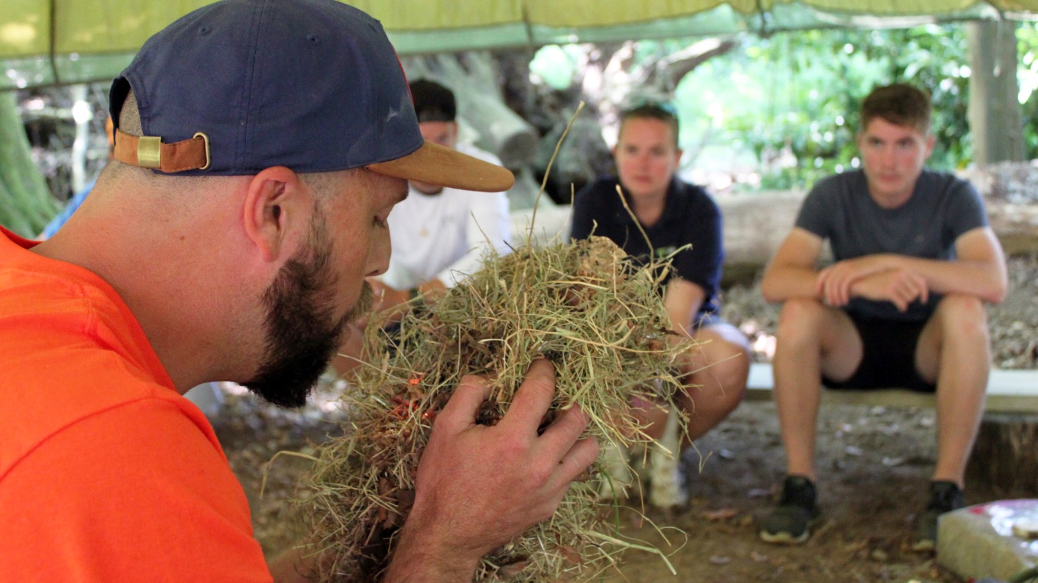 Man in cap demonstrates fire starting with dry grass to seated group in outdoor tent.