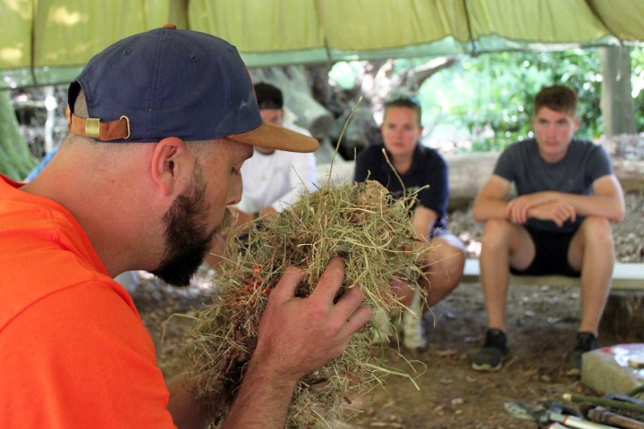 Man in cap demonstrates fire starting with dry grass to seated group in outdoor tent.