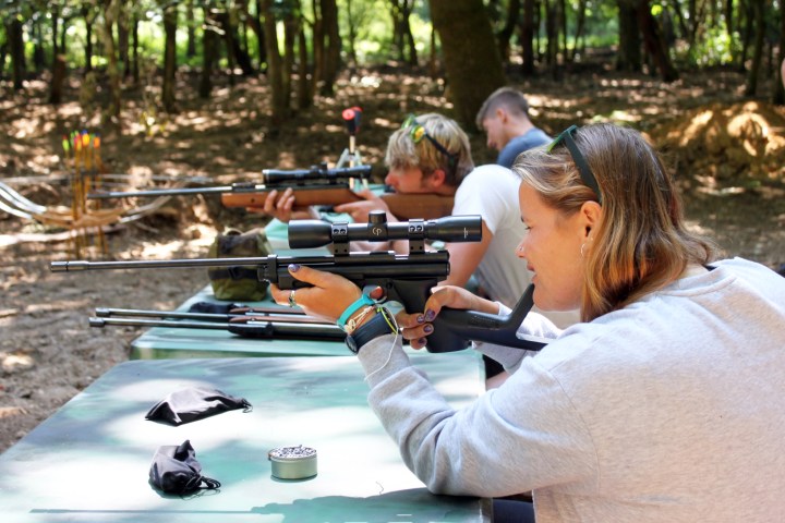 People at an outdoor range target shooting with rifles in a forest setting.