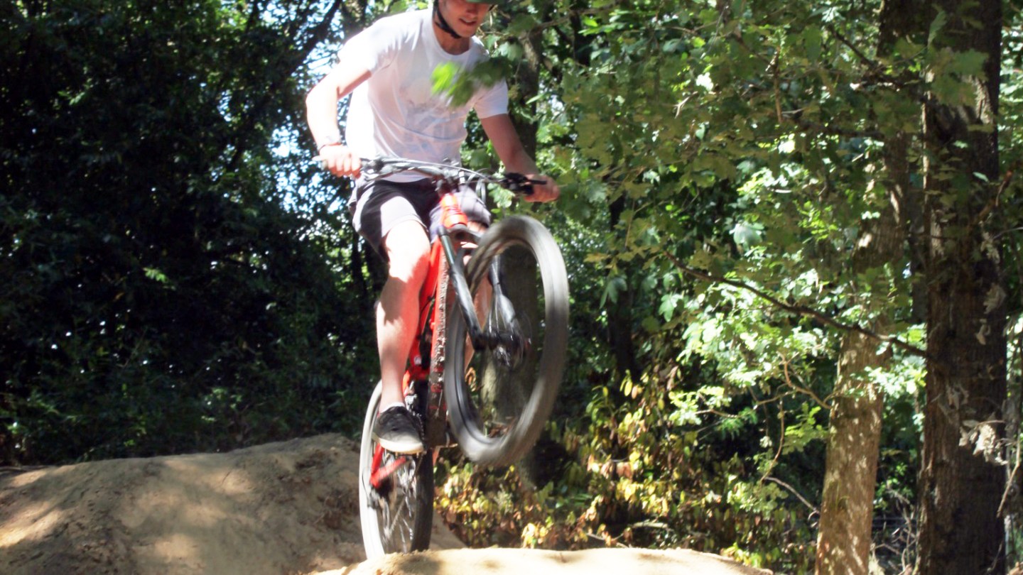 Person mountain biking over a dirt jump in a forested area.