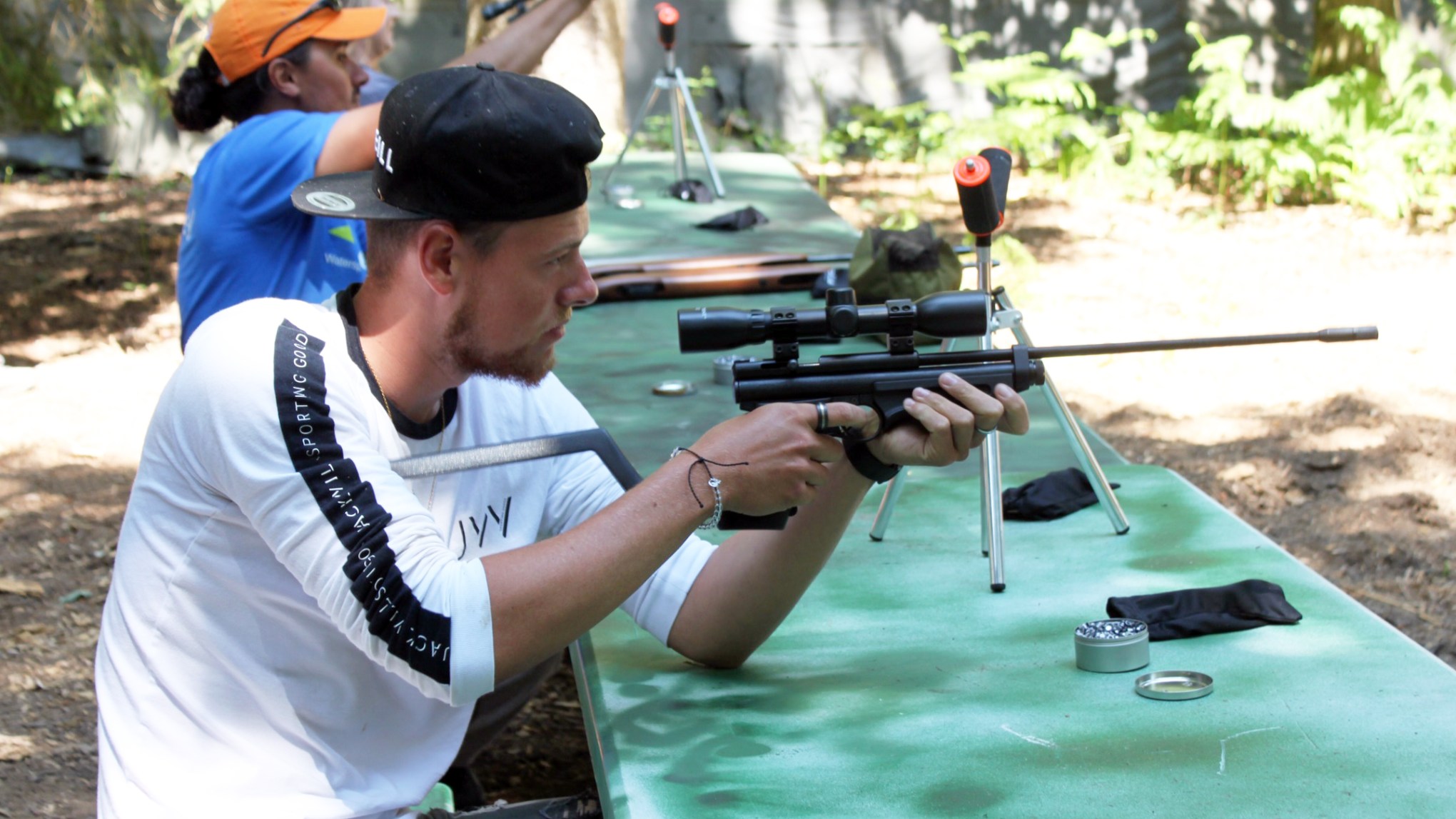 Two people aim rifles at a target range outdoors, with focus on the person in foreground.