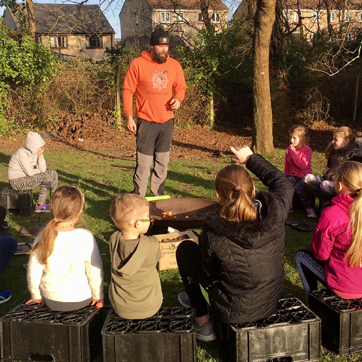 Children seated outdoors listening to two adults in orange shirts, with trees and grass around.