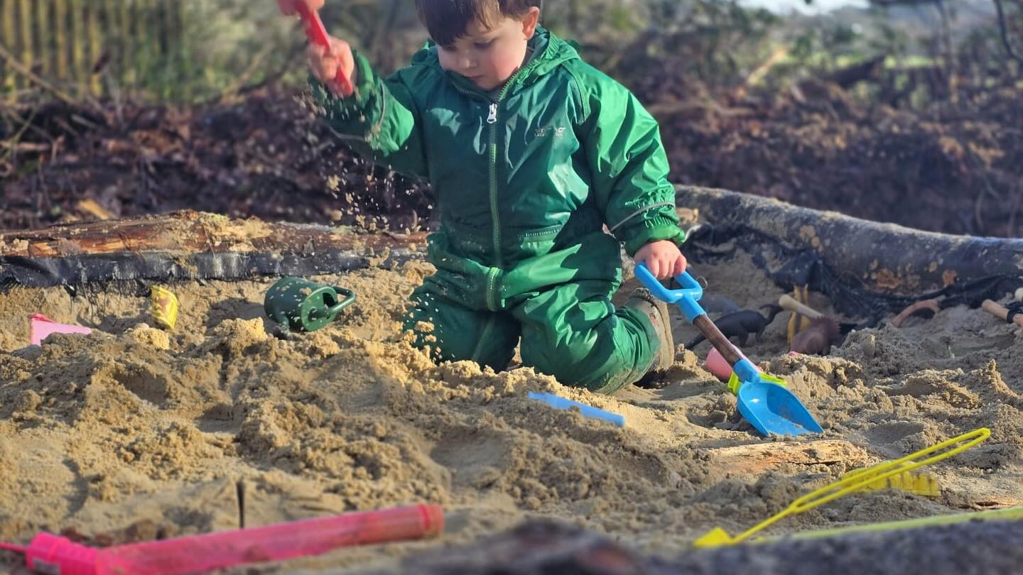 Child in green plays in a sandbox with colorful toys, under a tree.