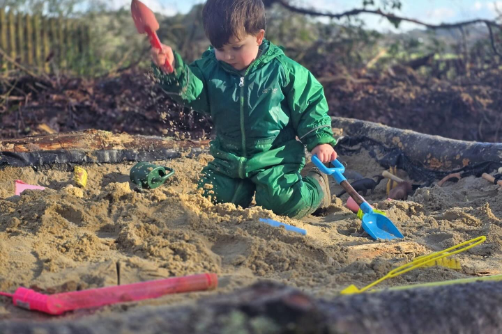 Child in green plays in a sandbox with colorful toys, under a tree.