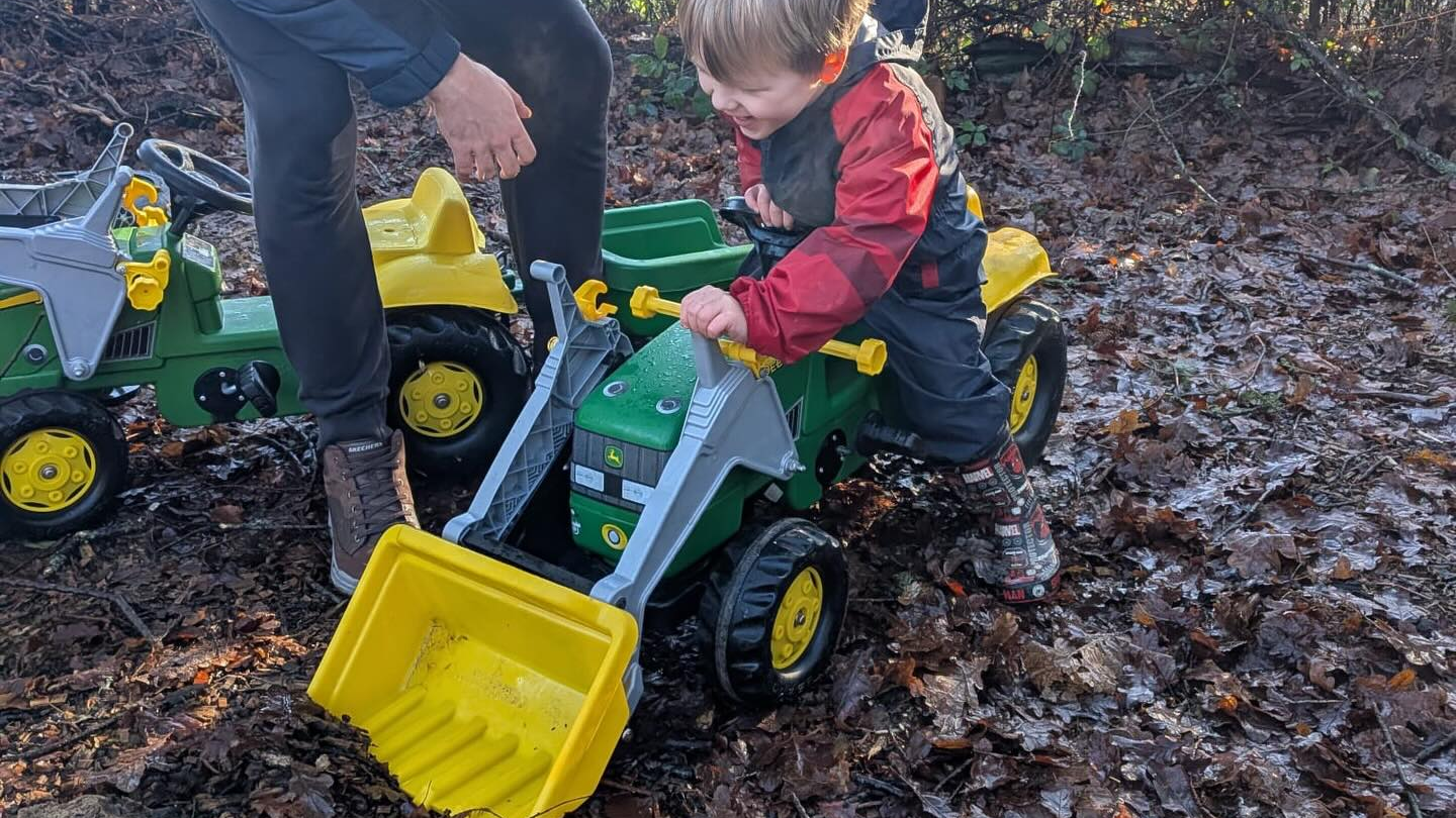 Adult helps child on toy tractor with yellow scoop, surrounded by leaves.