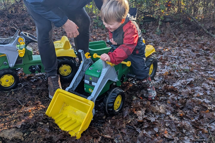 Adult helps child on toy tractor with yellow scoop, surrounded by leaves.