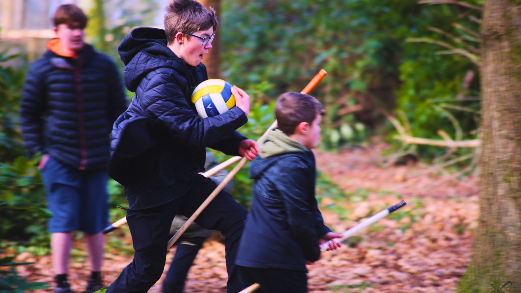 Children playing in a forest, one holding a volleyball, others with sticks, wearing jackets.