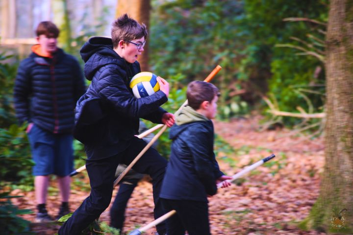 Children playing in a forest, one holding a volleyball, others with sticks, wearing jackets.