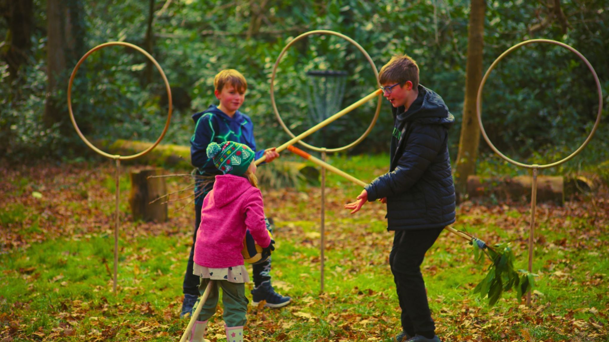Children playing with brooms in a forest, near large hoops on stands.