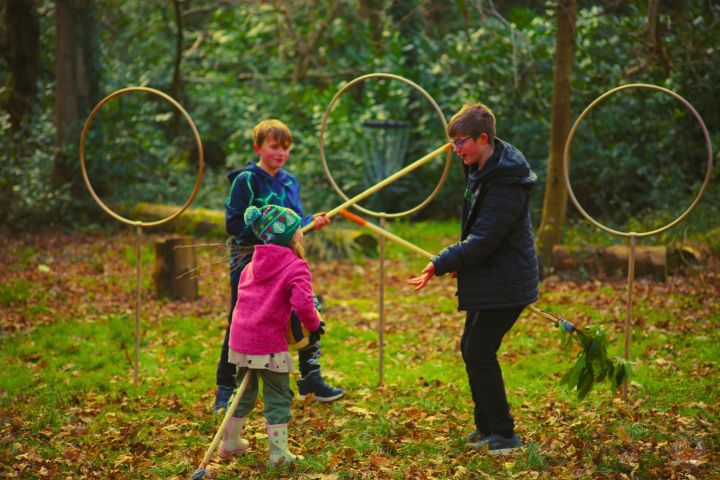 Children playing with brooms in a forest, near large hoops on stands.
