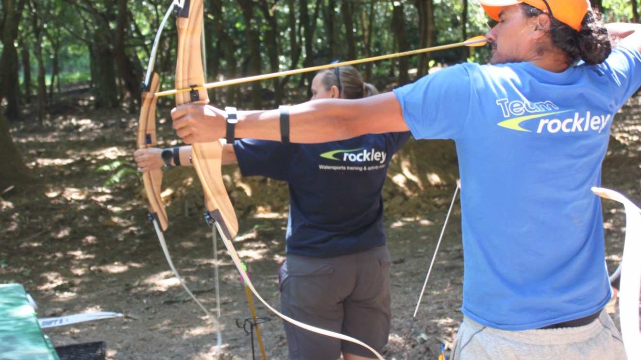 Two people practicing archery in a wooded area, drawing bows with arrows aimed ahead.