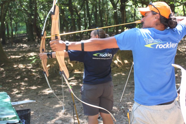 Two people practicing archery in a wooded area, drawing bows with arrows aimed ahead.