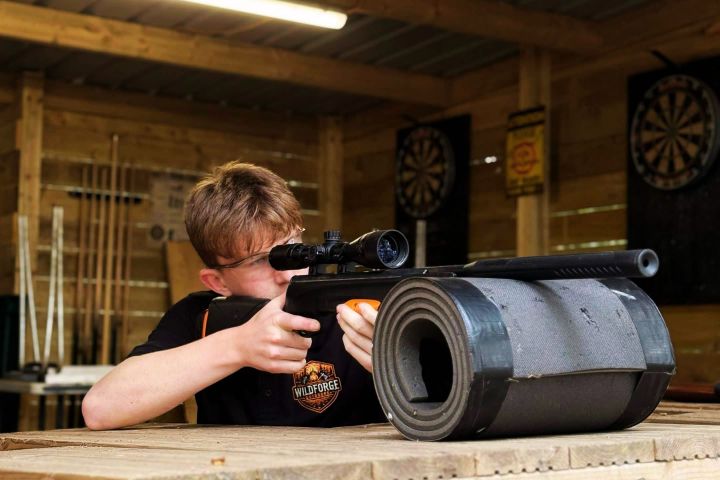 Person aiming a rifle indoors with a dartboard in the background.