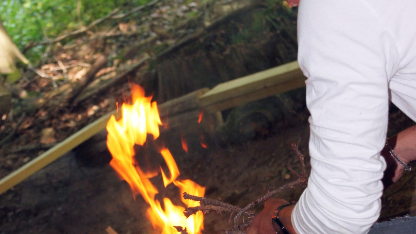 Person in a white shirt tending to a campfire in a forest setting.