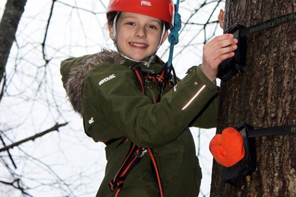 Person climbing a tree in a harness and red helmet, smiling.