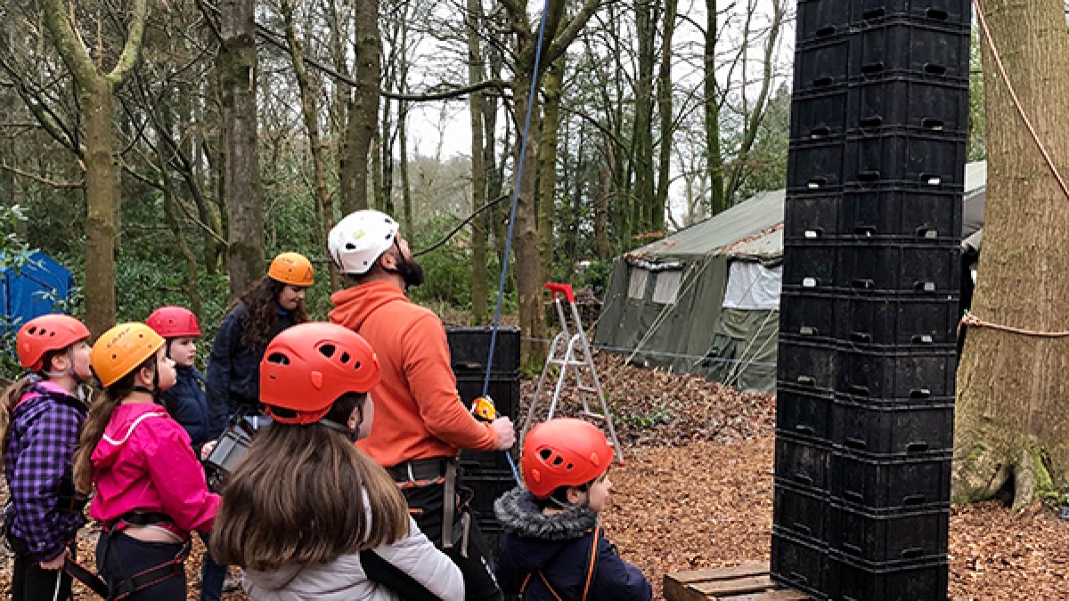 Group of people wearing helmets watch a climber stacking crates in a wooded area.