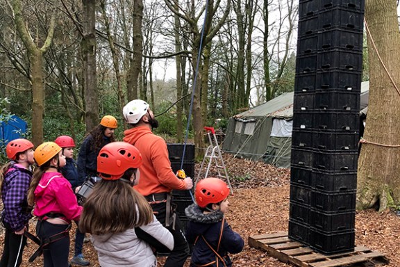 Group of people wearing helmets watch a climber stacking crates in a wooded area.