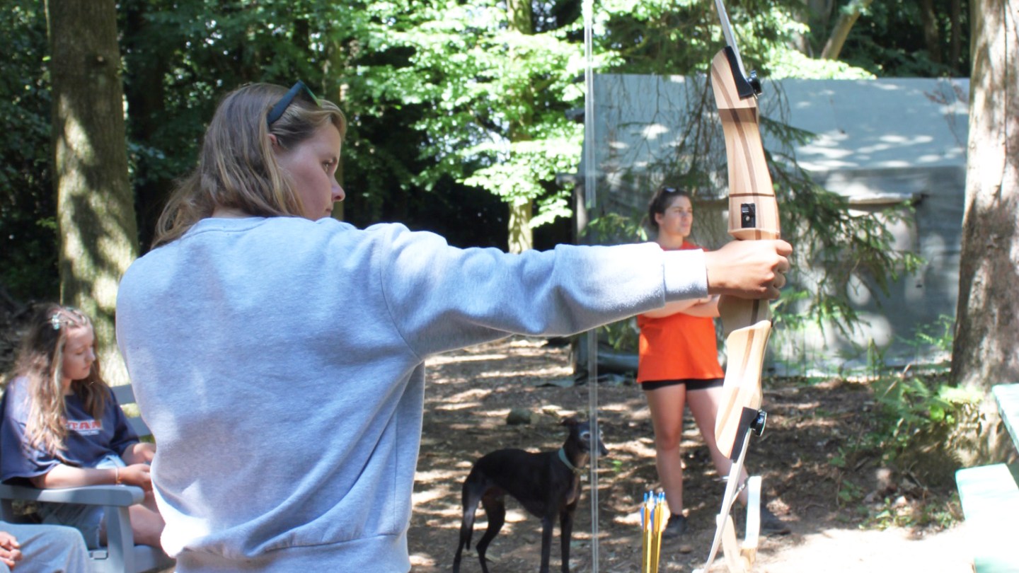 Person in gray shirt aiming a bow in a wooded area, with others and a dog in the background.