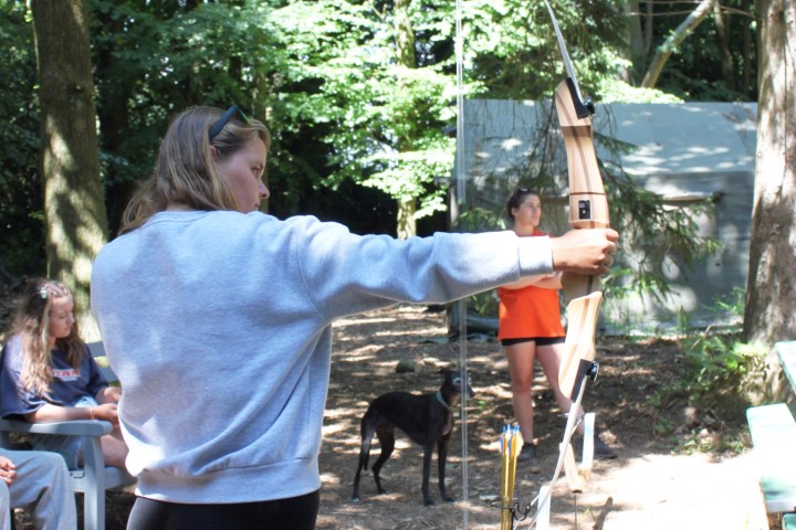 Person in gray shirt aiming a bow in a wooded area, with others and a dog in the background.