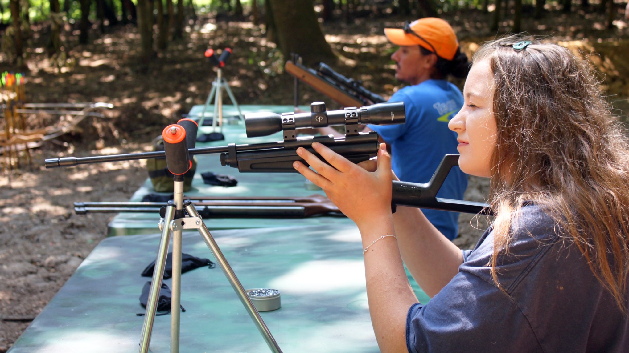 Two people aiming rifles at a shooting range with trees in the background.