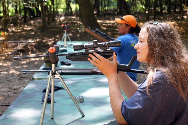 Two people aiming rifles at a shooting range with trees in the background.