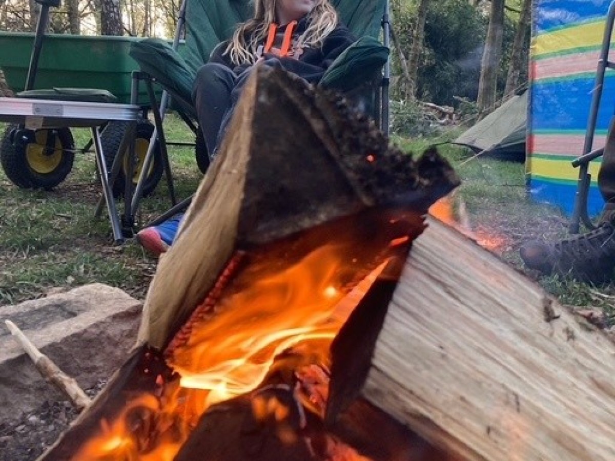 Person sitting by a campfire in a forest, with trees in the background.