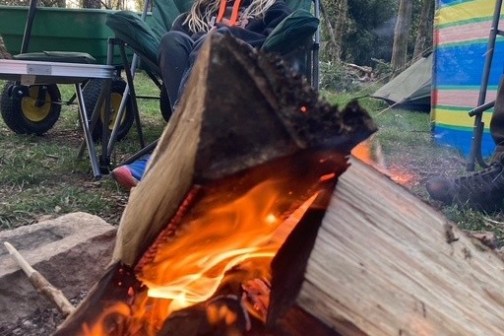 Person sitting by a campfire in a forest, with trees in the background.