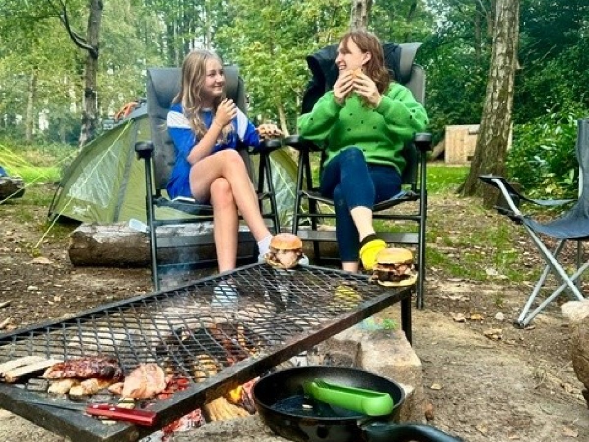 Two people eating by a campfire with a grill in a wooded area.