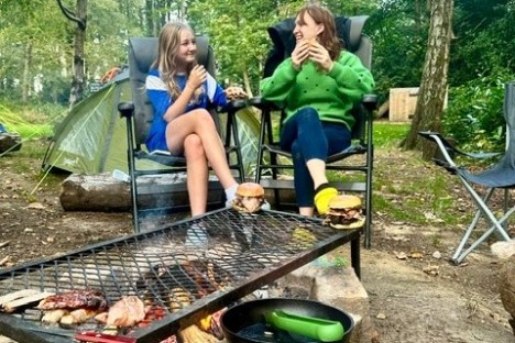 Two people eating by a campfire with a grill in a wooded area.