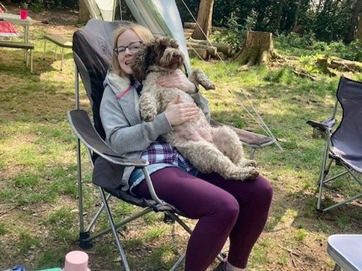 Person sitting in a camping chair holding a dog in a forest setting.