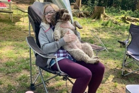 Person sitting in a camping chair holding a dog in a forest setting.