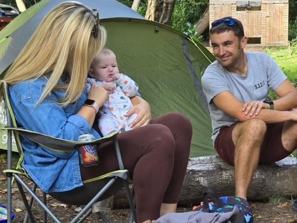 Two adults with a baby sitting outside a tent in a wooded area.