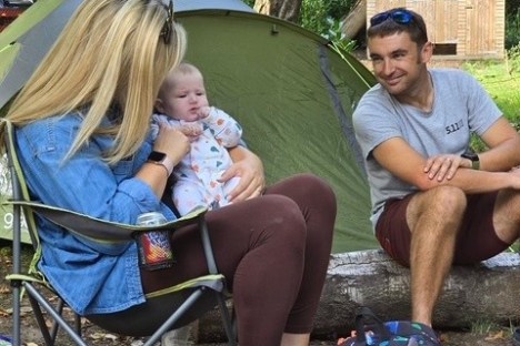 Two adults with a baby sitting outside a tent in a wooded area.