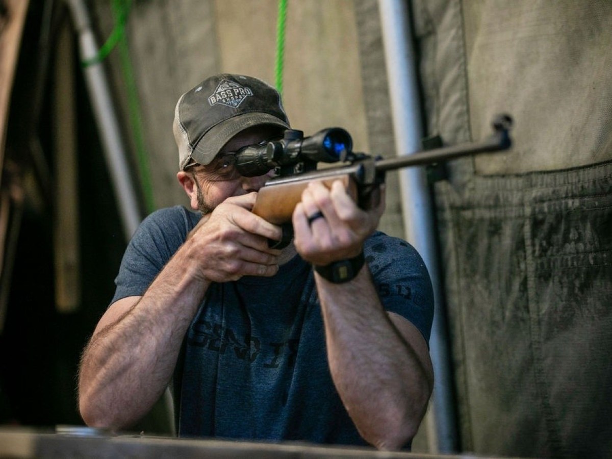 Person aiming rifle indoors with a scope.