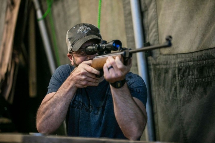 Person aiming rifle indoors with a scope.