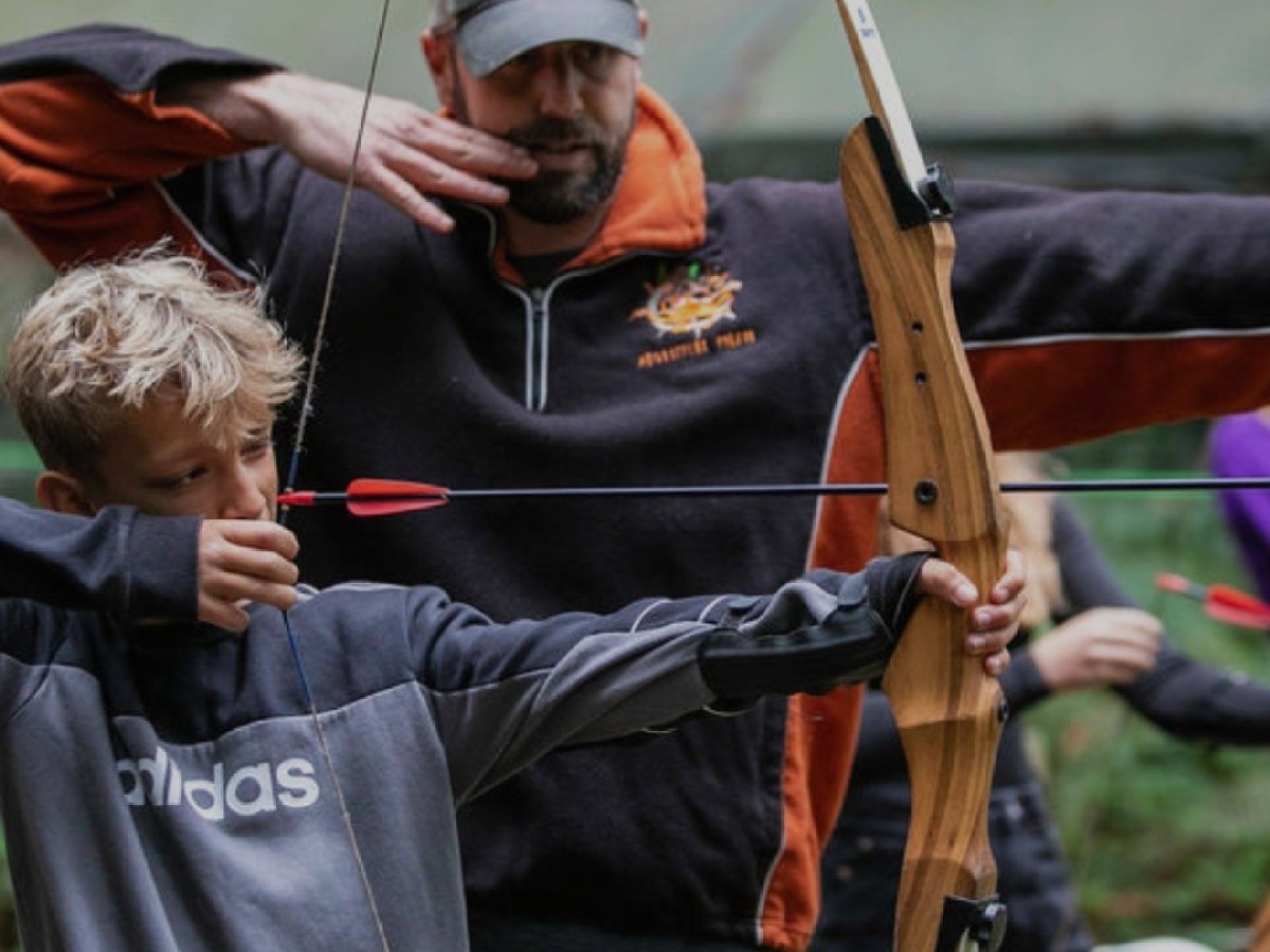 Man guides boy aiming bow and arrow in outdoor setting.