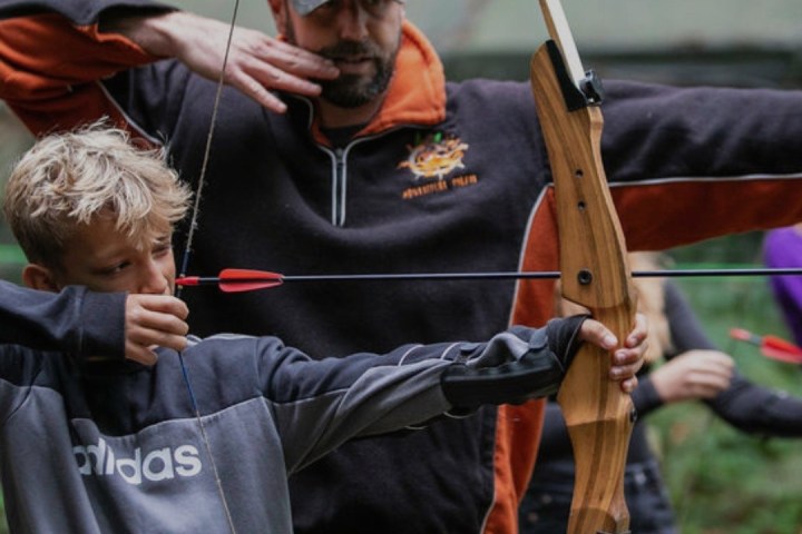Man guides boy aiming bow and arrow in outdoor setting.