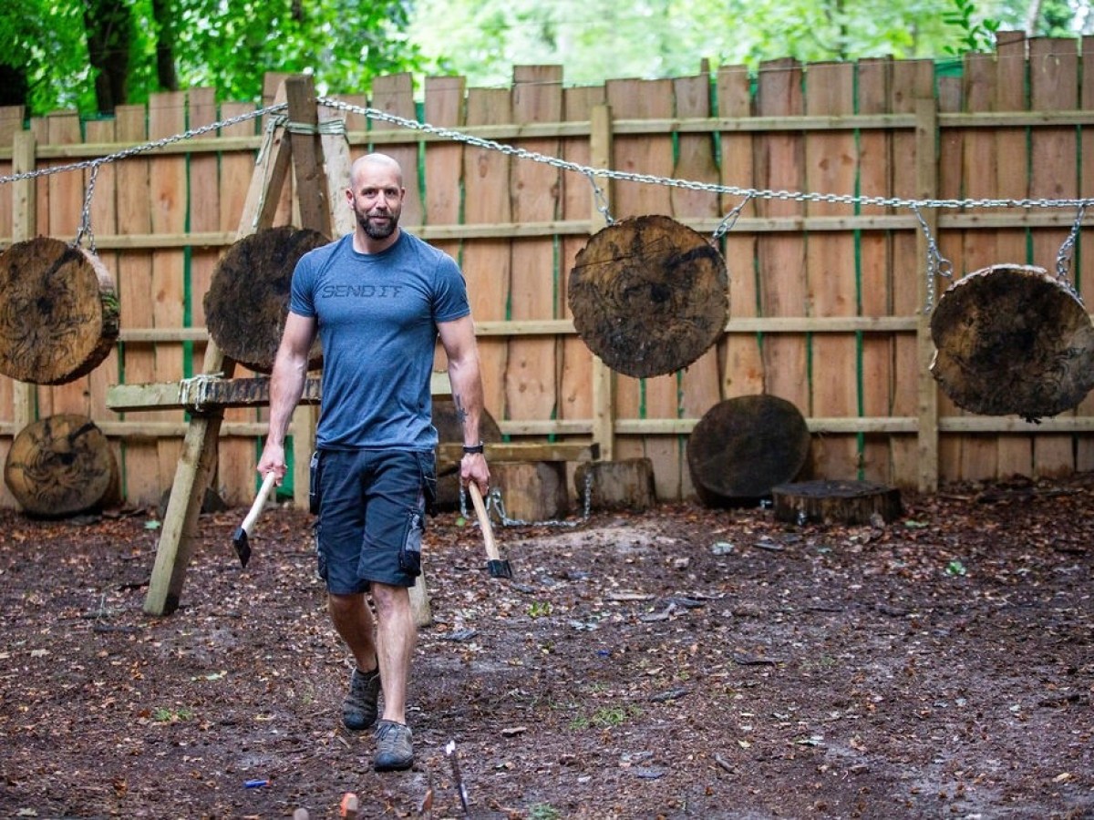 Man carrying axes in front of wooden targets in an outdoor area.
