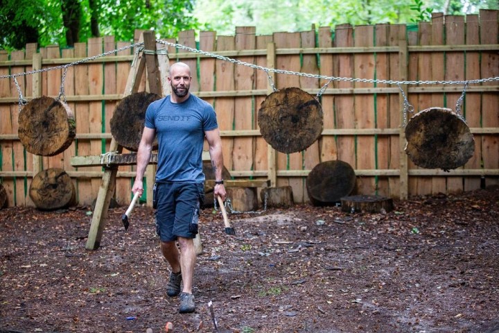 Man carrying axes in front of wooden targets in an outdoor area.