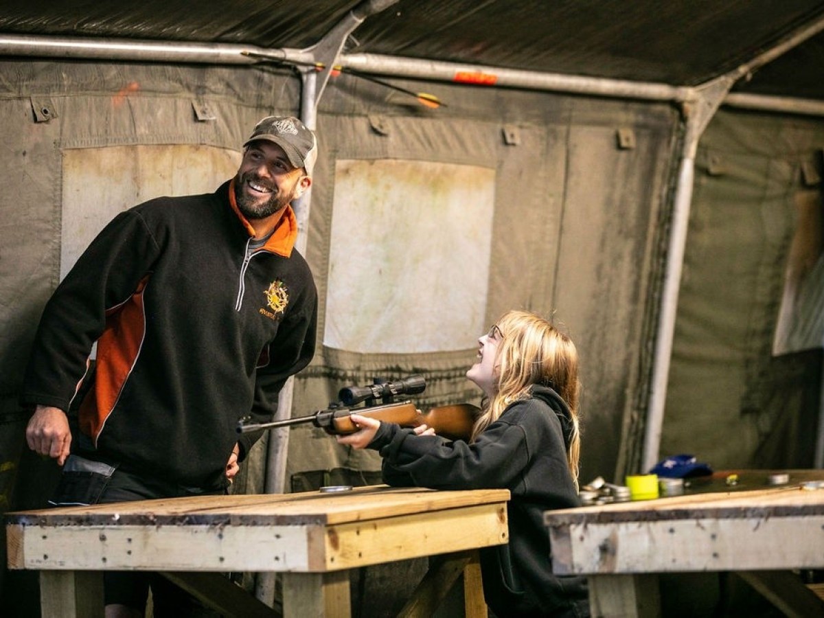 Man watches girl aiming a rifle at a shooting range.